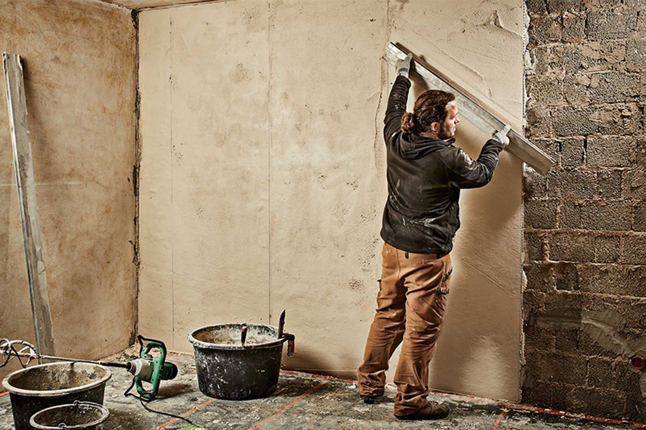 Handwerker glättet Putz an einer Wand. Eimer, Werkzeuge und eine lange Richtlatte sind im Raum verteilt.