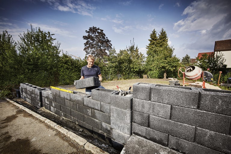 Un homme construit un mur en parpaings dans le jardin, avec une bétonnière en arrière-plan.