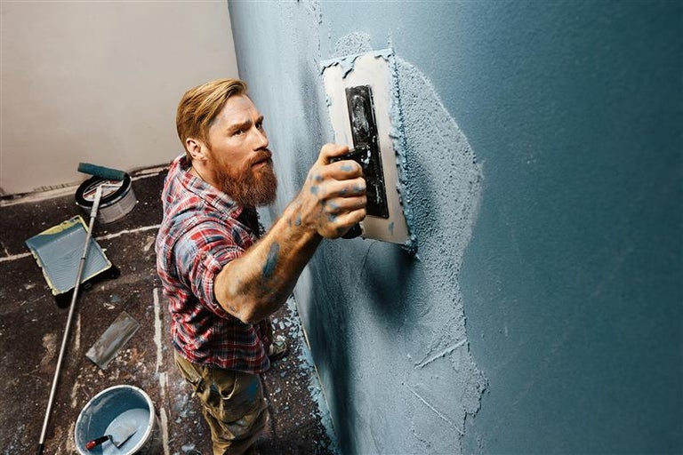 Un homme enduit un mur avec une taloche dans une pièce contenant du matériel de peinture.