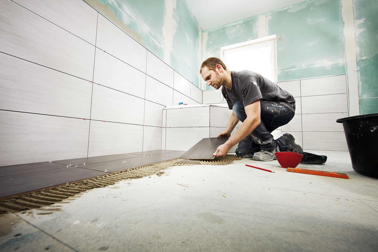 Ein Mann verlegt Fliesen im Badezimmer, wobei er Fliesenkleber und Abstandhalter verwendet.