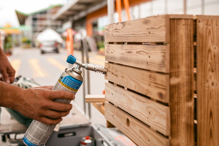 Person bearbeitet eine Holzkiste mit einem Gasbrenner.