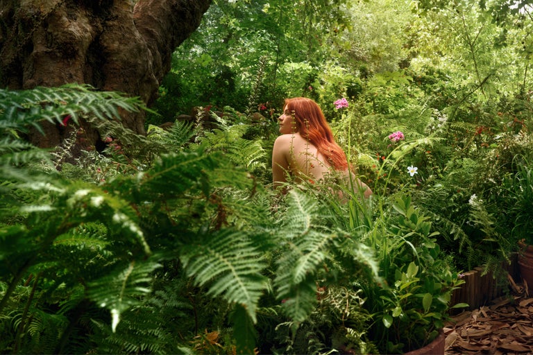 Frau mit roten Haaren sitzt inmitten eines üppigen Gartens mit Farnen und blühenden Pflanzen