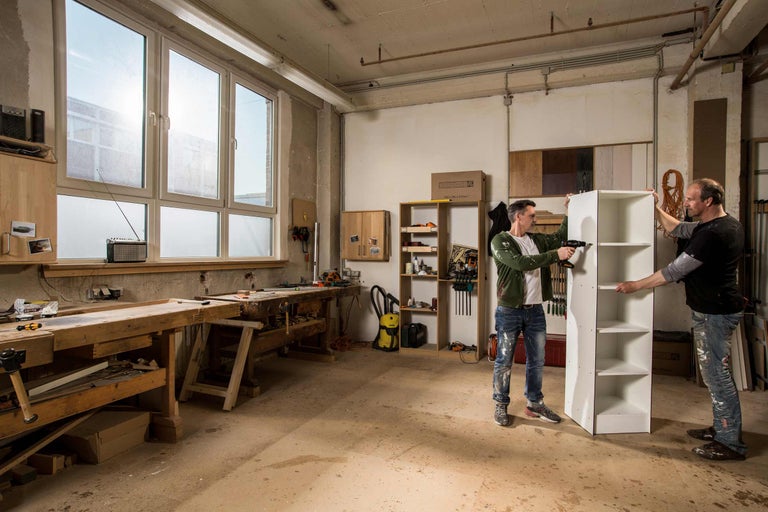 Deux personnes montent une étagère dans un atelier. Des établis et des outils sont visibles à l'arrière-plan.