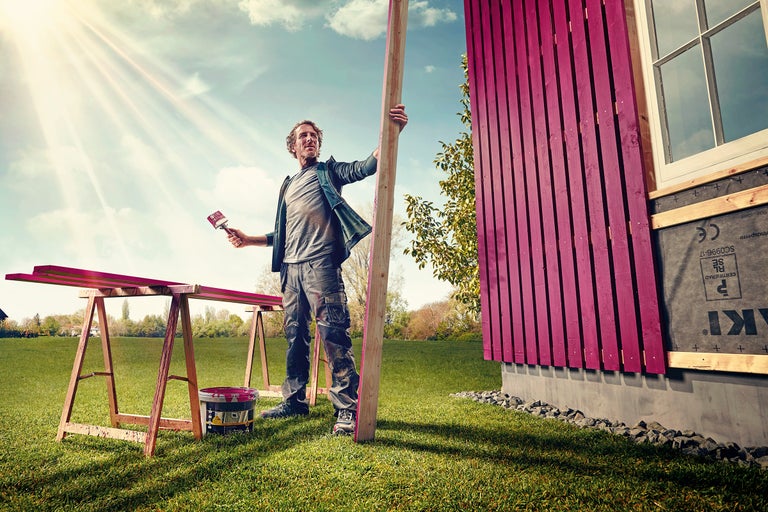 Mann streicht Holzbretter und Hauswand mit Farbe im Garten