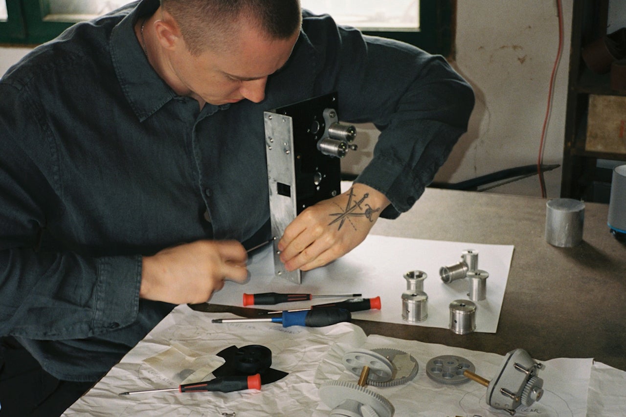 Ein Handwerker montiert mit Schraubendrehern präzise mechanische Metallteile und Zahnräder an einem Arbeitstisch in einer Werkstatt.