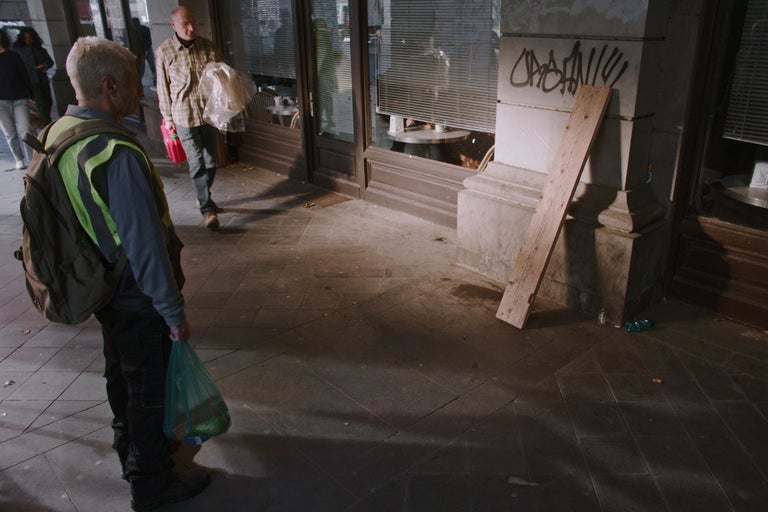 Städtische Aussenszene mit einem Holzbrett an einer Steinfassade und einem Mann in Warnweste im Vordergrund.