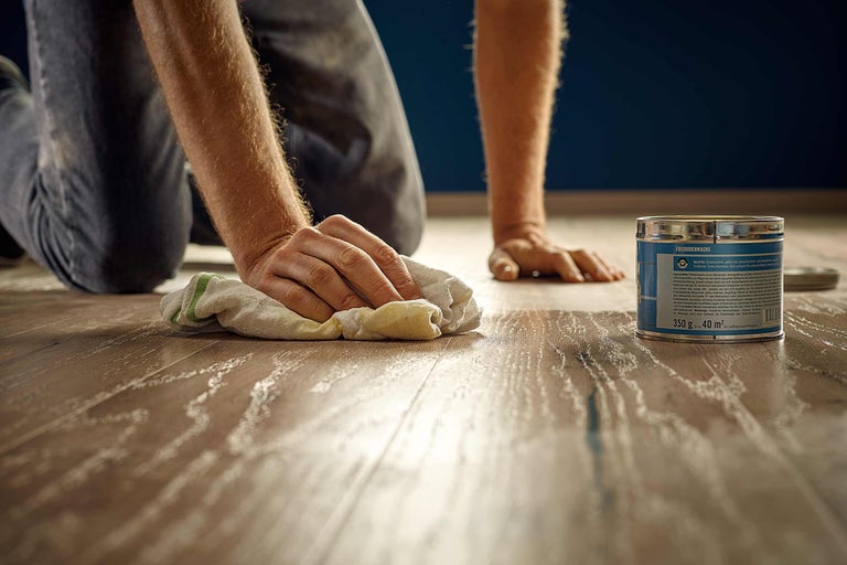 Une personne applique de la cire pour parquet sur un plancher en bois à l'aide d'un chiffon, une boîte de cire pour parquet est visible en arrière-plan.