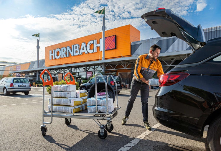 Ein Mitarbeiter von Hornbach belädt einen Einkaufswagen mit Baumaterialien und räumt diese in ein Auto vor dem Hornbach Markt.