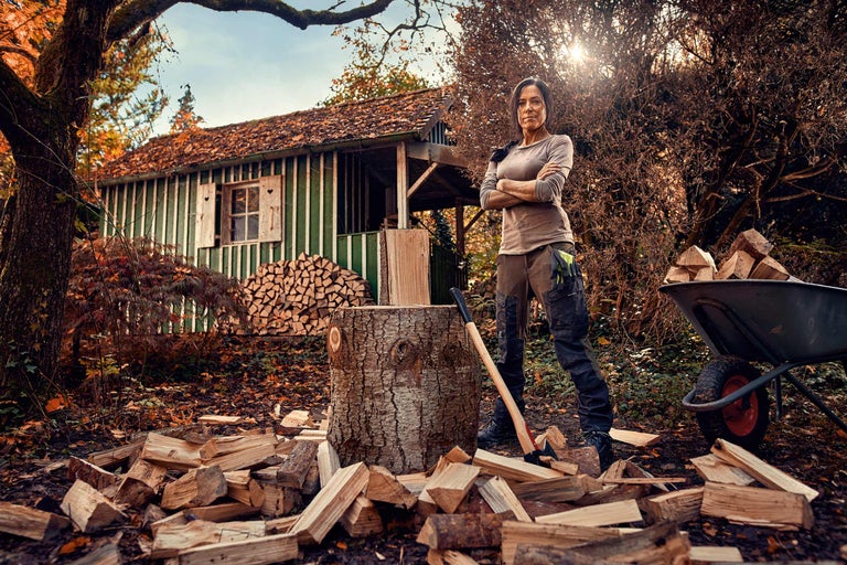 Frau mit Holzspaltaxt, Holzscheiten, Schubkarre und Holzhütte im Hintergrund