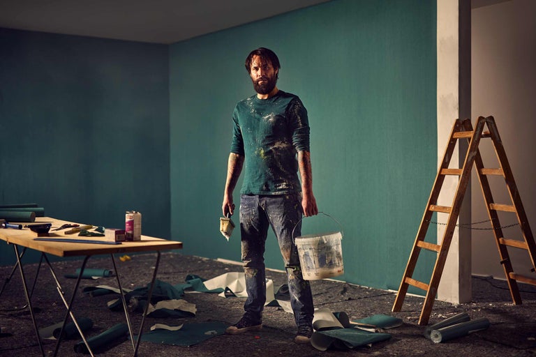 Mann streicht Wand mit Farbe im Innenraum, inklusive Leiter und Malerwerkzeug