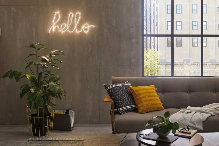 Wohnzimmer mit Sofa, Kissen, Zimmerpflanze im gelben Topf und leuchtendem Schriftzug Hello an der Wand.