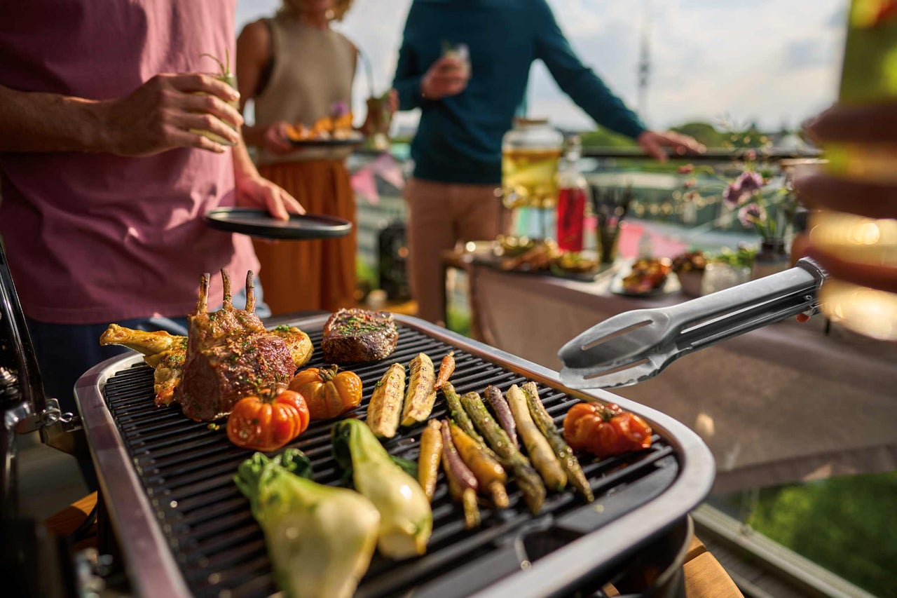 Szene mit einem Grill, auf dem Fleisch, Gemüse und Grillzange liegen, während im Hintergrund Personen stehen
