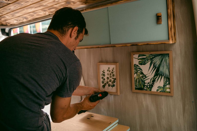 Mann befestigt Bilderrahmen mit einem Akkubohrer an der Wand in einem Fahrzeug.