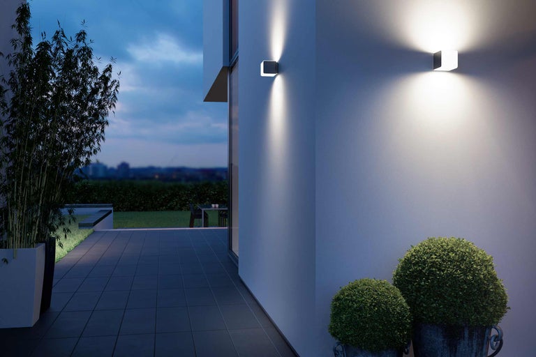 Beleuchtete Aussenansicht mit zwei quadratischen Wandleuchten, Bepflanzung und Terrassenbereich.
