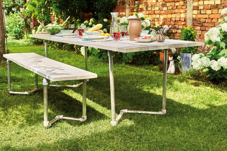 Scène de jardin avec table à manger et banc en bois et métal sur pelouse, dressée avec nourriture et boissons