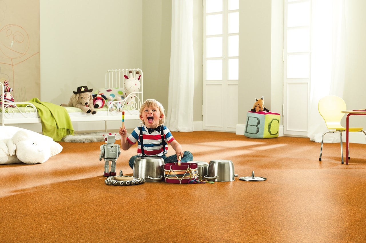 Helles Kinderzimmer mit Korkboden, in dem ein Kind mit Töpfen und einem Spielzeugroboter spielt. Im Hintergrund sind ein Bett und eine Sitzecke zu sehen.