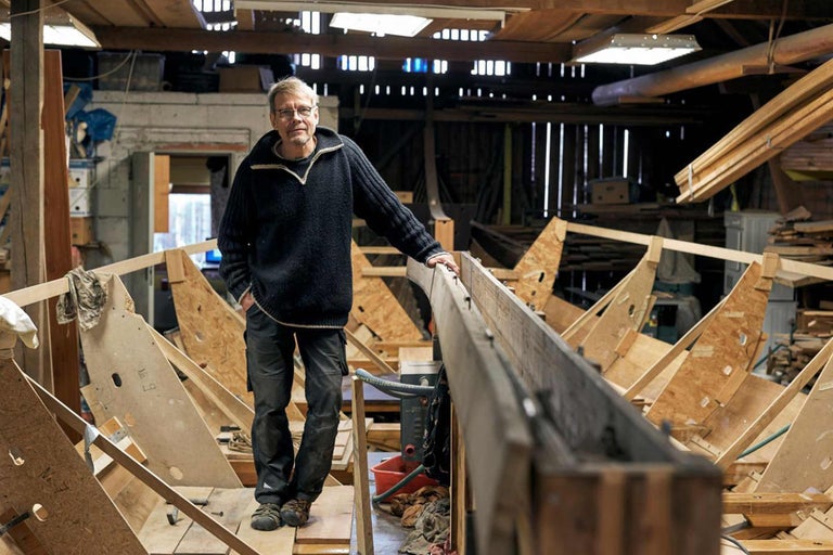 Mann steht in einer Werkstatt vor einem unfertigen Holzboot.