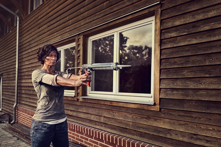 Frau benutzt eine Kartuschenpistole an einem Haus mit Holzfassade und weißen Fensterrahmen