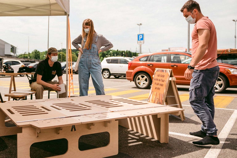 Szene im Freien mit drei Personen, einem Holztisch und Autos auf einem Parkplatz.