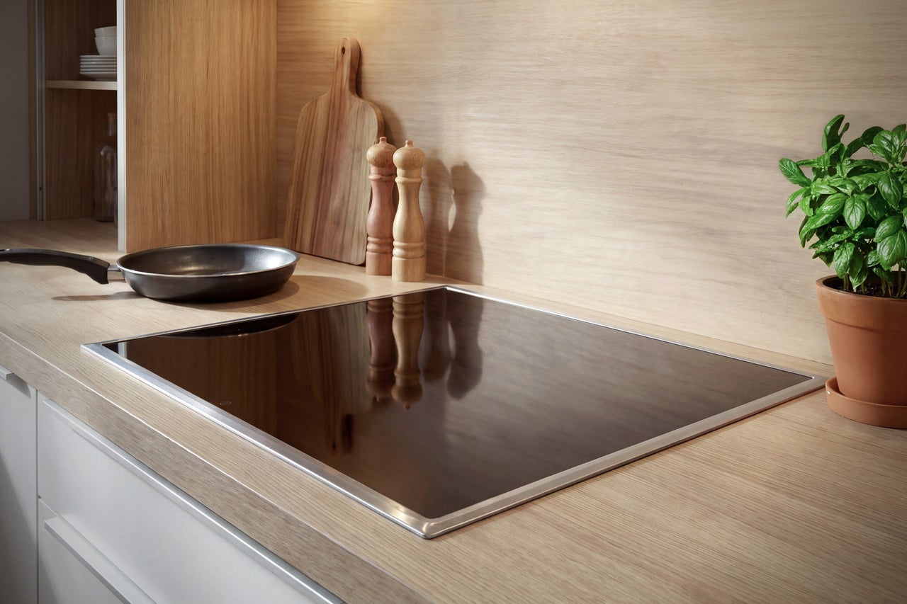 Plaque de cuisson à induction encastrable avec cadre en acier inoxydable dans une cuisine au décor bois clair, avec poêle, moulins et basilic.