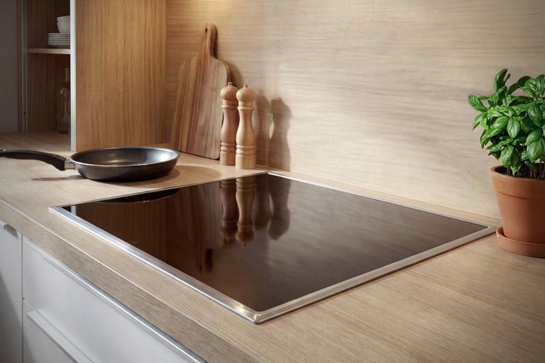 Plaque de cuisson à induction encastrable avec cadre en acier inoxydable dans une cuisine au décor bois clair, avec poêle, moulins et basilic.