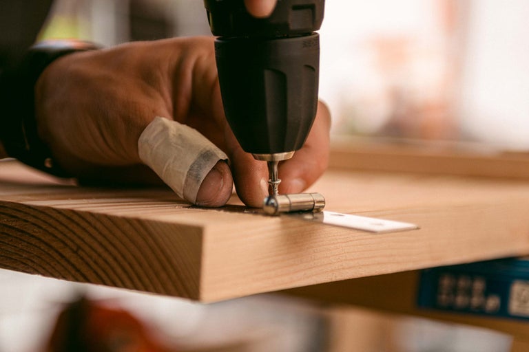 Eine Person befestigt mit einem Akkubohrer ein Scharnier an einem Holzbrett.