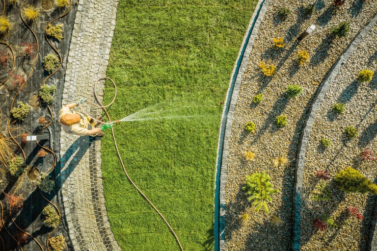 Luftaufnahme einer Person, die einen Rasen mit einem Wasserschlauch bewässert, neben einem mit Kies und Pflanzen bedeckten Bereich