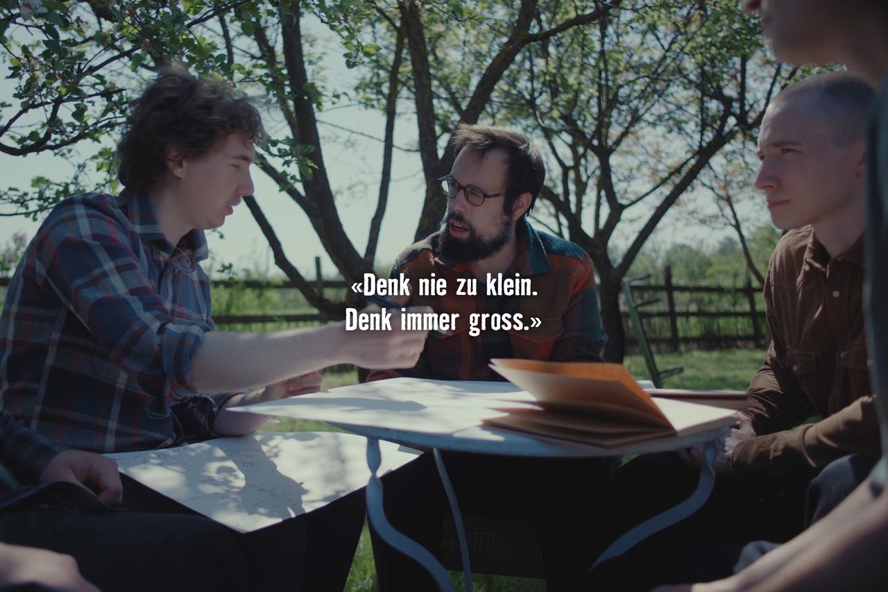 Drei Männer sitzen im Freien an einem Tisch und besprechen Planungen mit Papier und Stift unter einem Baum.