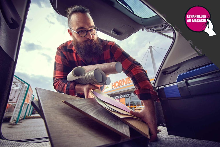 Un homme charge des matériaux de construction dans le coffre d'une voiture, notamment des rouleaux de papier peint, du stratifié et des nuanciers devant un magasin de bricolage Hornbach.