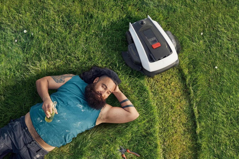 Un homme barbu est allongé sur une pelouse à côté d'un robot tondeuse et tient une bouteille à la main.