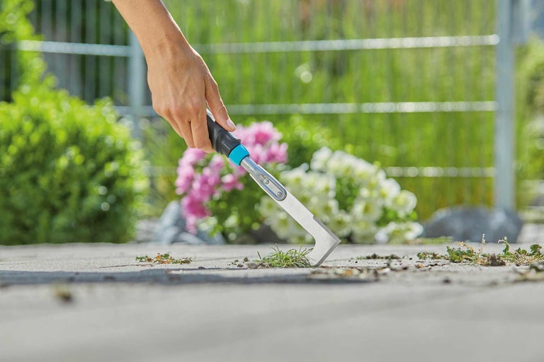 Une main tient un grattoir à joints pour enlever les mauvaises herbes entre les pavés.