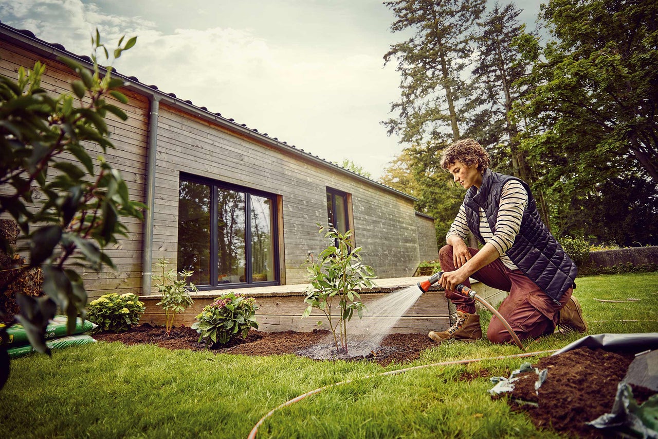 Eine Person bewässert junge Pflanzen in einem Garten mit einem Haus im Hintergrund.
