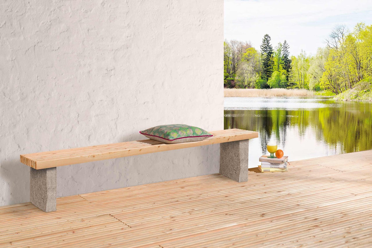 Holzbank mit Steinfüssen, Kissen, Getränk und Büchern auf einer Holzterrasse mit Seeblick