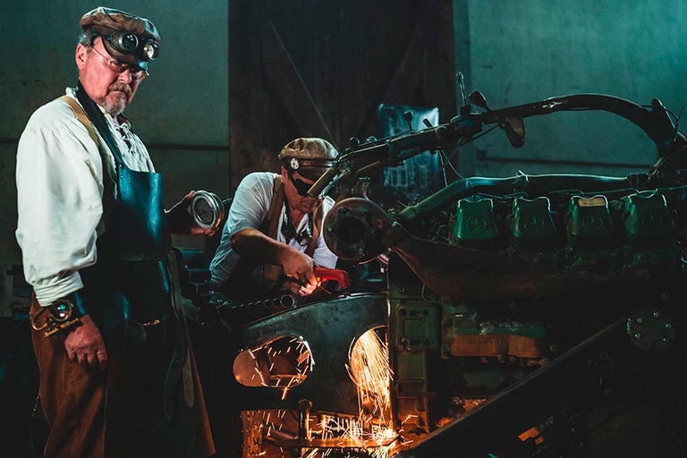 Deux artisans travaillent sur une ancienne machine avec une meuleuse et des lunettes de protection
