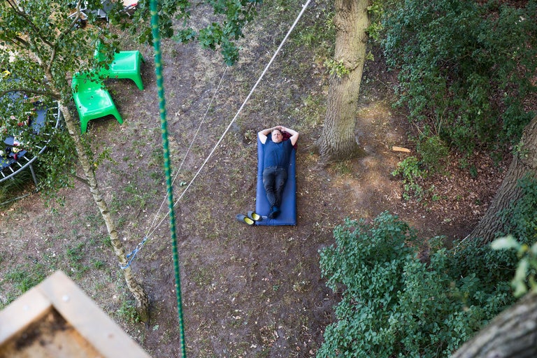 Draufsicht auf einen Mann, der auf einer blauen Isomatte im Garten liegt, umgeben von Bäumen, Rasen, grünen Stühlen und Seilen.