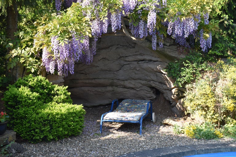 Scène avec une chaise longue dans le jardin, entourée de plantes et d'un mur en pierre avec des glycines