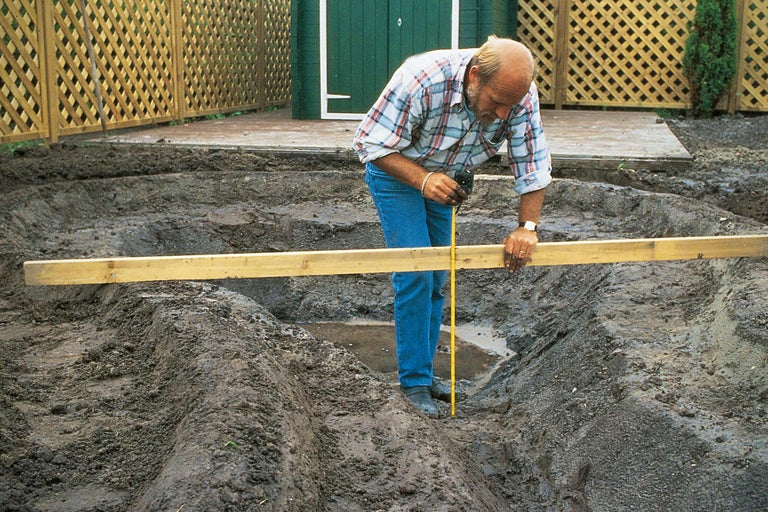 Ein Mann misst mit einem Messband die Tiefe einer Ausgrabung für einen Gartenteich