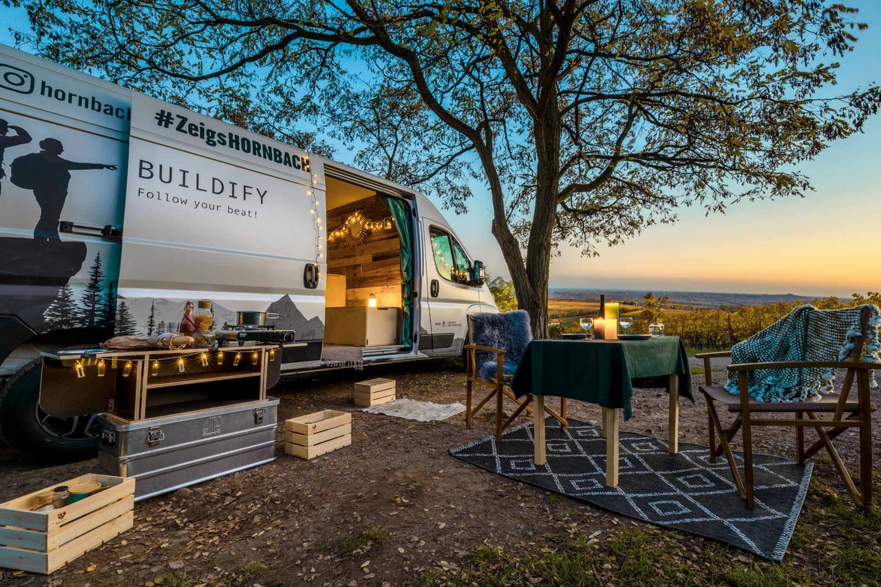 Scène avec un van aménagé, une table de camping et des chaises de camping en extérieur.