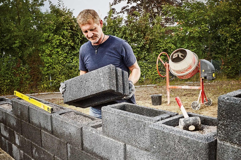 Mann baut eine Mauer mit Betonsteinen, unterstützt durch eine Wasserwaage und einen Gummihammer, mit einem Betonmischer im Hintergrund.