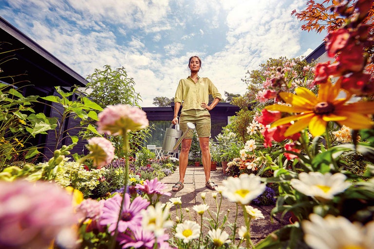 Femme avec arrosoir se tient dans un jardin coloré.