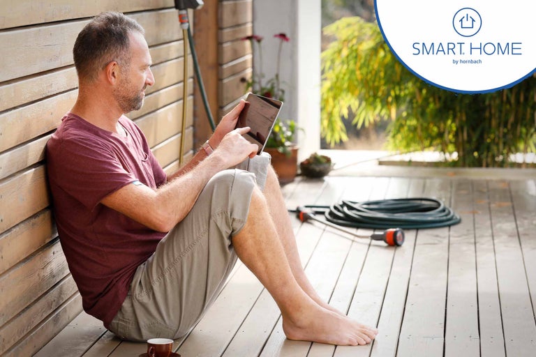 Un homme est assis avec une tablette sur une terrasse en bois ; tuyau d'arrosage en arrière-plan ; Logo Hornbach Smart Home