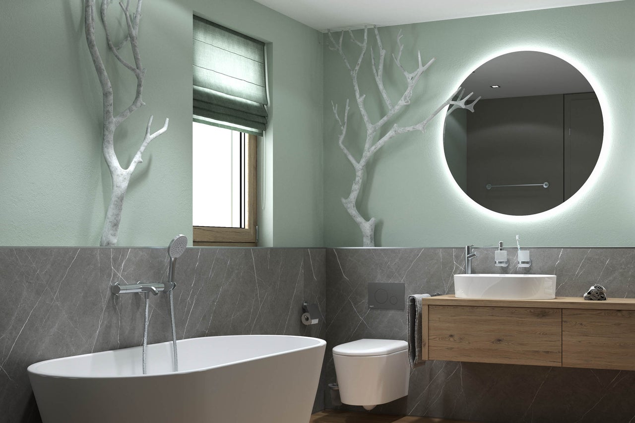 Salle de bains moderne avec baignoire en îlot, toilettes suspendues et meuble sous-vasque en bois. Grand miroir rond rétroéclairé et branches décoratives.