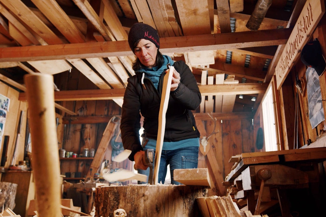 Eine Frau arbeitet mit einer Axt in einer Holzwerkstatt an einem Holzstück.