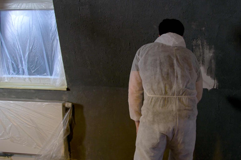 Personne en combinaison de protection devant un mur crépi avec fenêtre et radiateur masqués