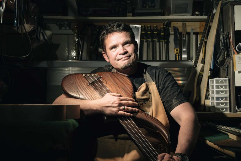 Mann im Handwerkerlook hält ein Holz-Saiteninstrument in seiner Werkstatt