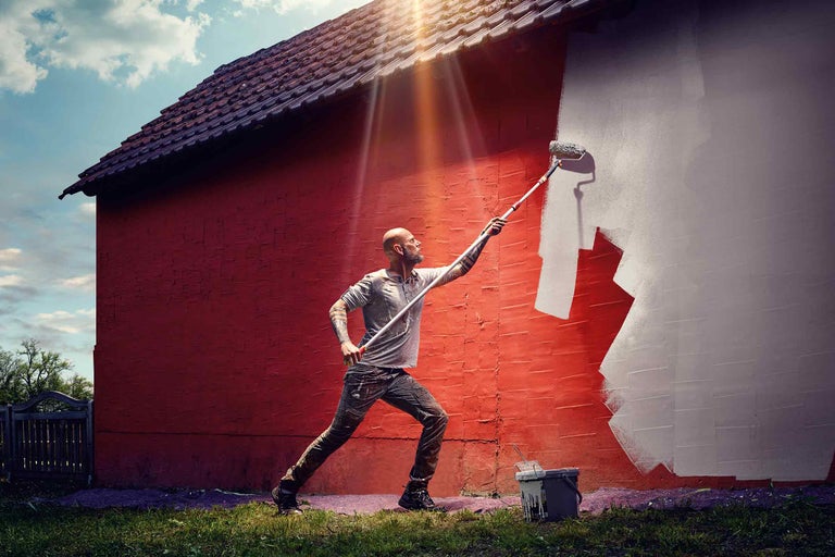 Un homme peint un mur de maison avec un rouleau à peinture et de la peinture.