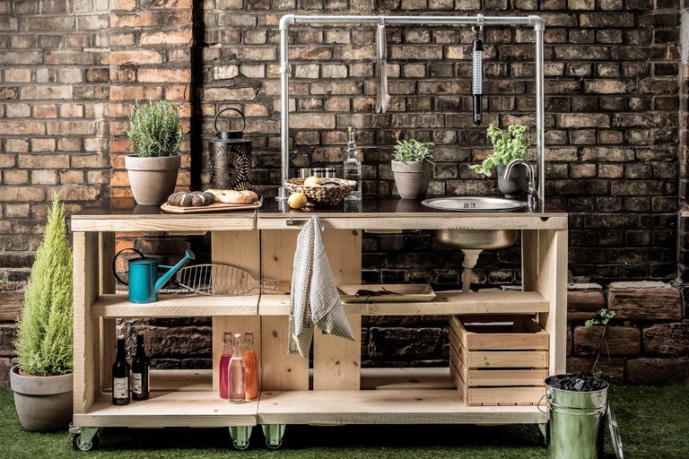 Cuisine extérieure avec structure en bois, évier, étagères et roulettes devant un mur de briques