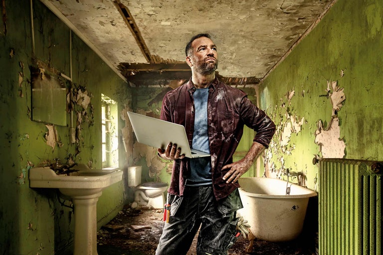 Artisan avec ordinateur portable dans une salle de bains à rénover avec baignoire, toilettes et lavabo