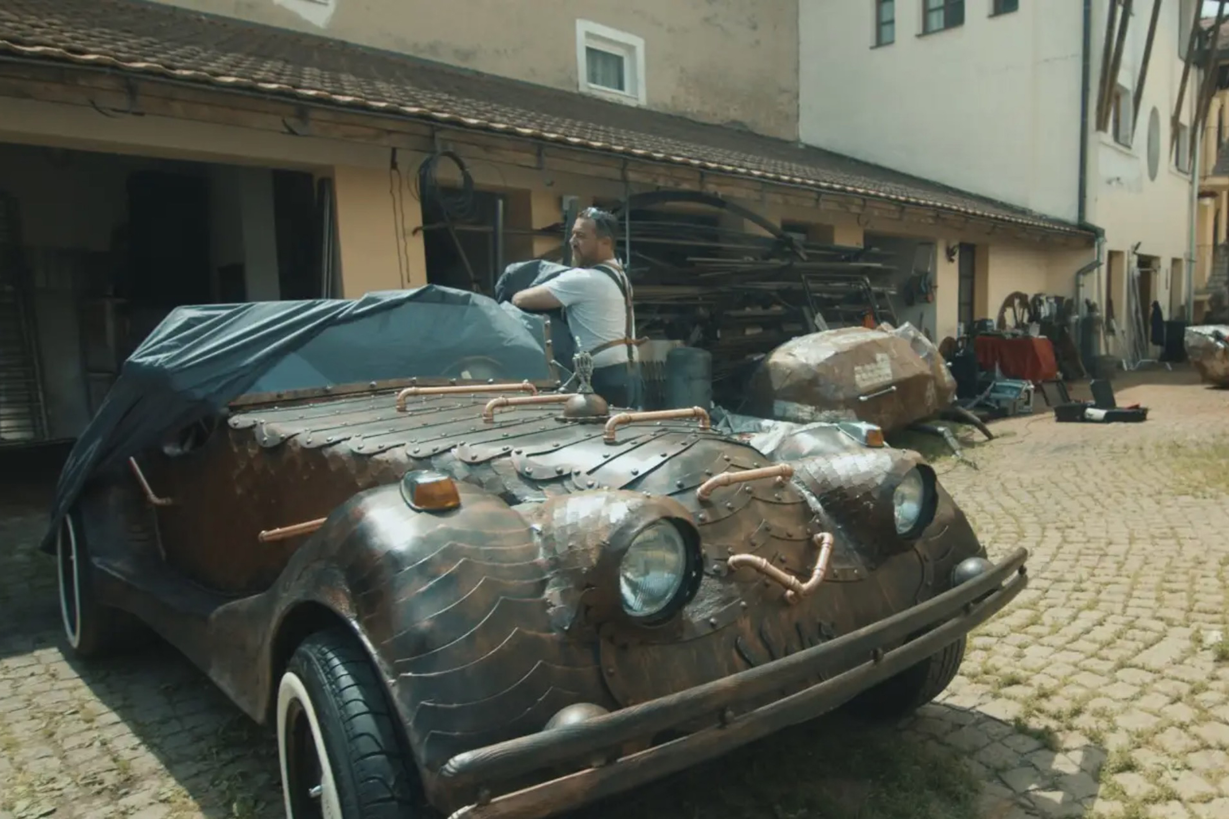 Une voiture personnalisée avec des détails en cuivre, partiellement recouverte d'une bâche, est garée à l'extérieur.