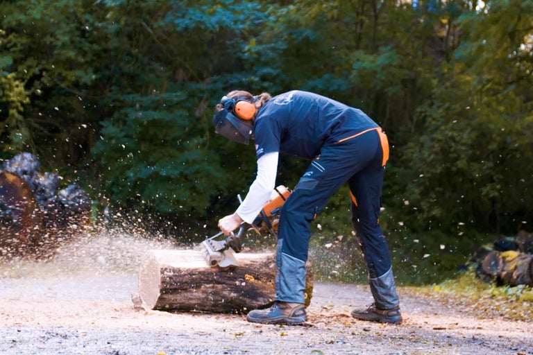 Une personne scie une bûche avec une tronçonneuse, portant des vêtements de protection et une protection auditive.